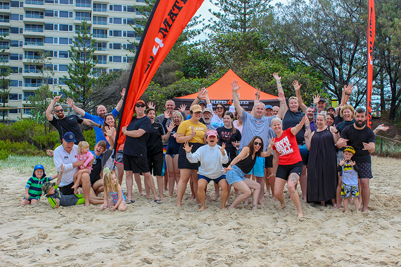 Group of people standing on the beach with Mates4Mates banners and gazebos promoting a fundraiser. 