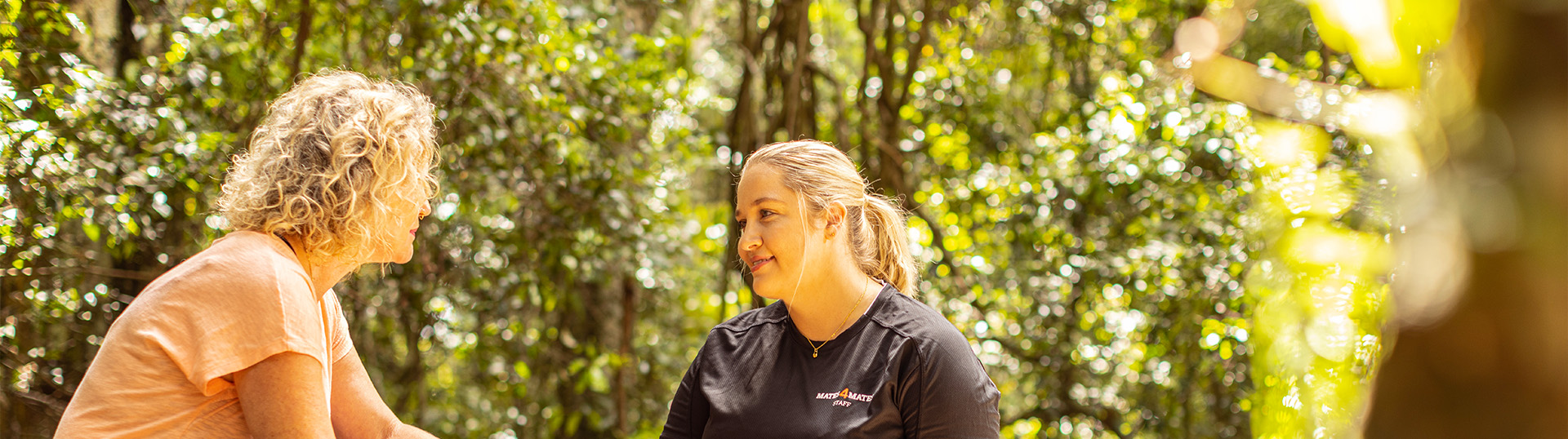 Mates4Mates staff member kneeling down talking with a female client who is sitting on a log outdoors with trees in the background.