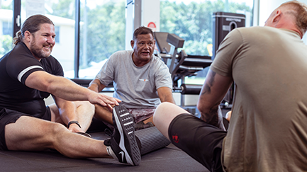 Three men sit on the floor of the gym and stretch their legs.
